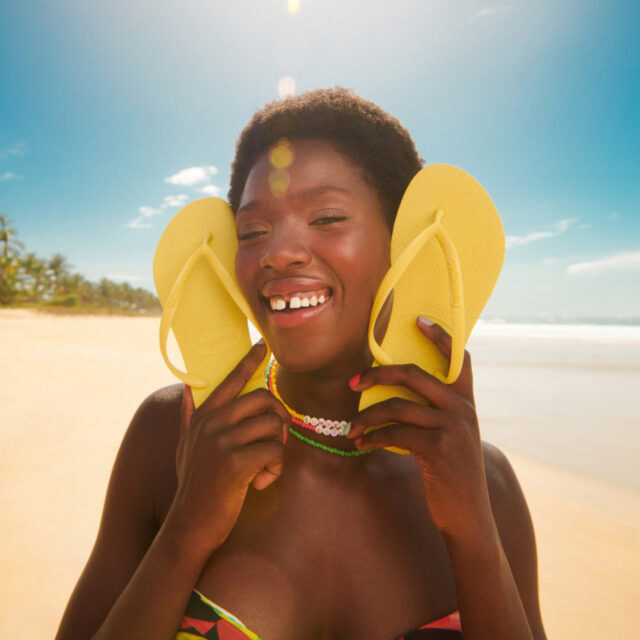 A woman holding yellow flip-flops by her cheeks smiles joyfully on a sunny beach.