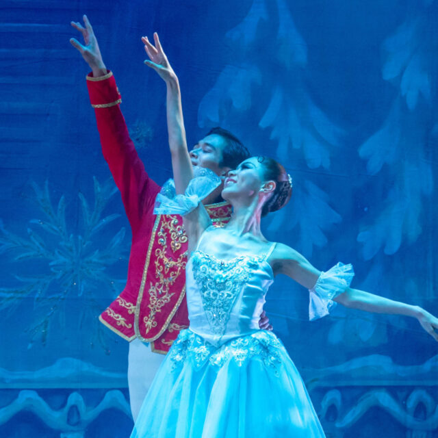 Two ballet dancers on stage, performing a pose; the male in a red jacket, the female in a blue tutu, with a blue scenic backdrop.
