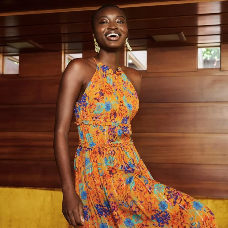 Person in an orange floral dress stands inside a wood-paneled room, smiling with one foot slightly raised.