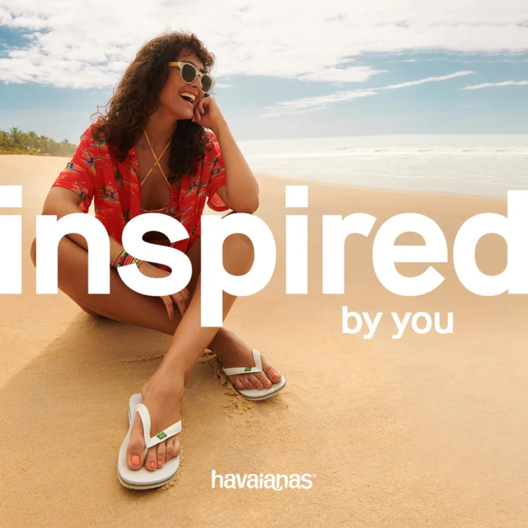 Woman in a red shirt and sunglasses sits on a sandy beach with white flip-flops nearby. Large text reads "inspired by you" with "havaianas" at the bottom.