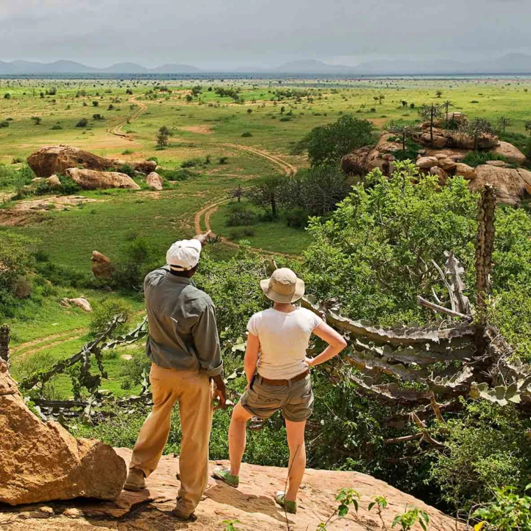 Two people wearing hats stand on a rocky ledge, overlooking a vast, green savanna landscape with distant mountains under a cloudy sky.