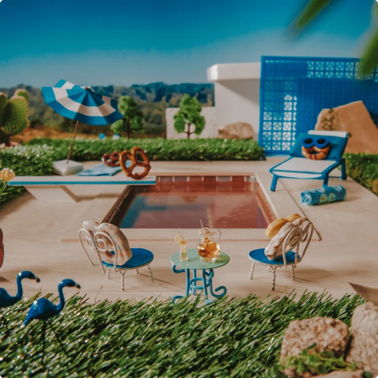 Whimsical scene of a poolside setting with pretzel figures sitting on chairs. A pool, umbrella, and lounge chair are in the background, surrounded by faux grass and decorative elements.