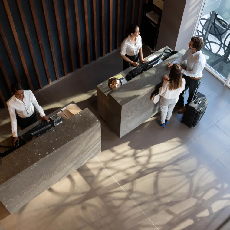 Hotel lobby reception with two people checking in at the front desk. Two staff members assist them while another staff member is at a separate desk. Suitcase beside the guests.