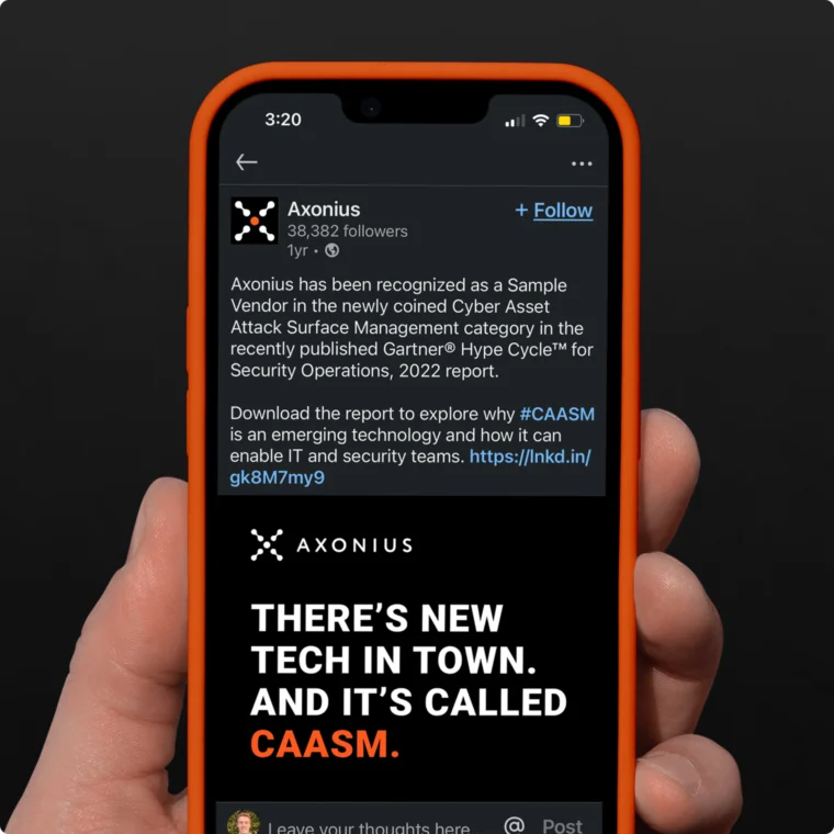 Hand holding a smartphone displaying a LinkedIn post from Axonius about CAASM recognition by Gartner, with a black background and white text saying "There's New Tech in Town. And It's Called CAASM.