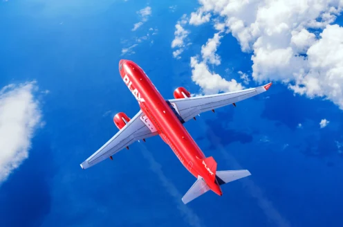 A red commercial airplane flies above a blue ocean with scattered white clouds in the sky.