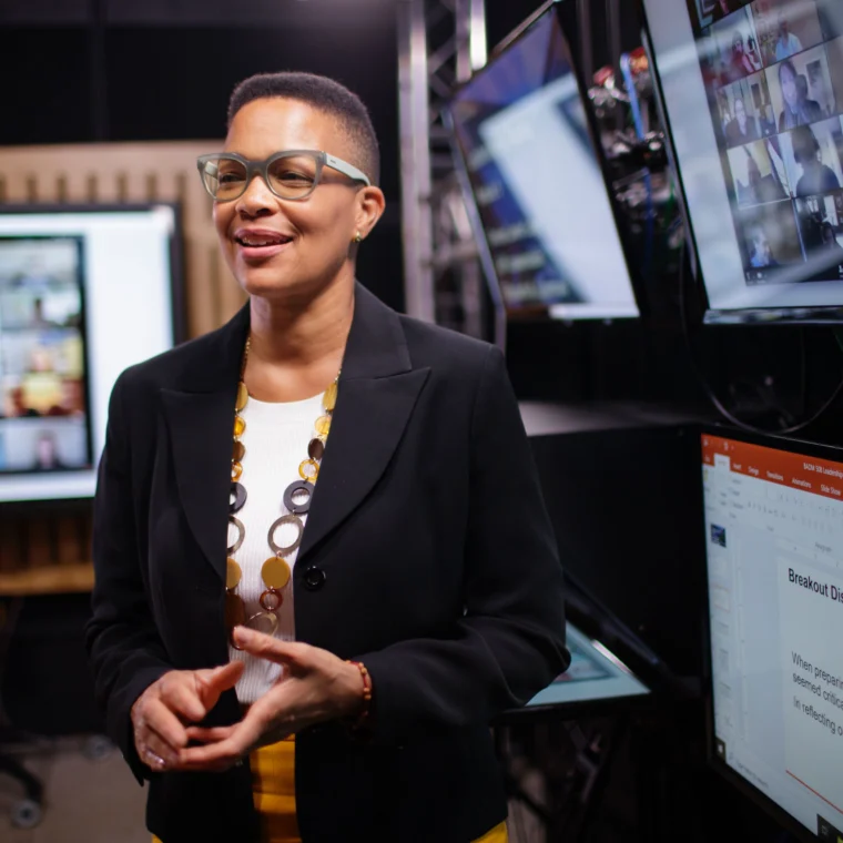 Person speaking in a room with multiple screens displaying virtual meeting participants and presentation slides.