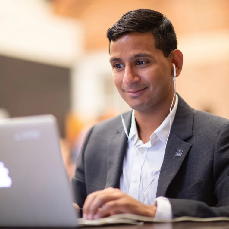 A man in a suit smiling while using a laptop, wearing earphones.