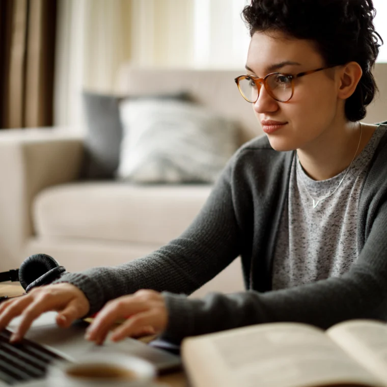 Person wearing glasses and a sweater sits on the floor using a laptop, with an open book nearby in a cozy living room setting.