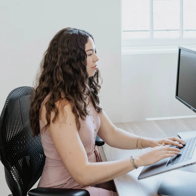 Person with long curly hair typing on a computer keyboard, seated at a wooden desk in a bright room with a window.
