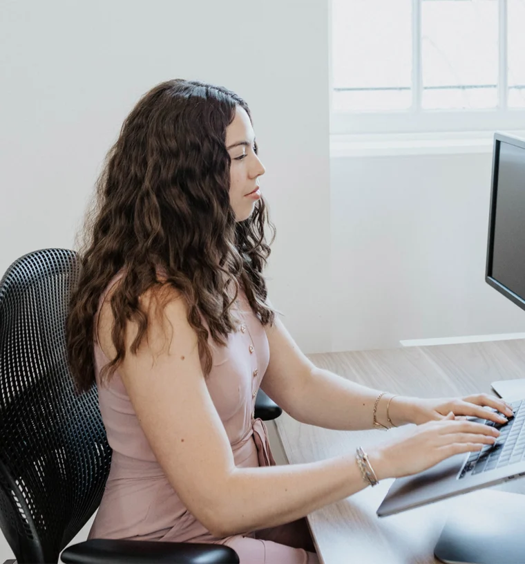 Person with long curly hair typing on a computer keyboard, seated at a wooden desk in a bright room with a window.
