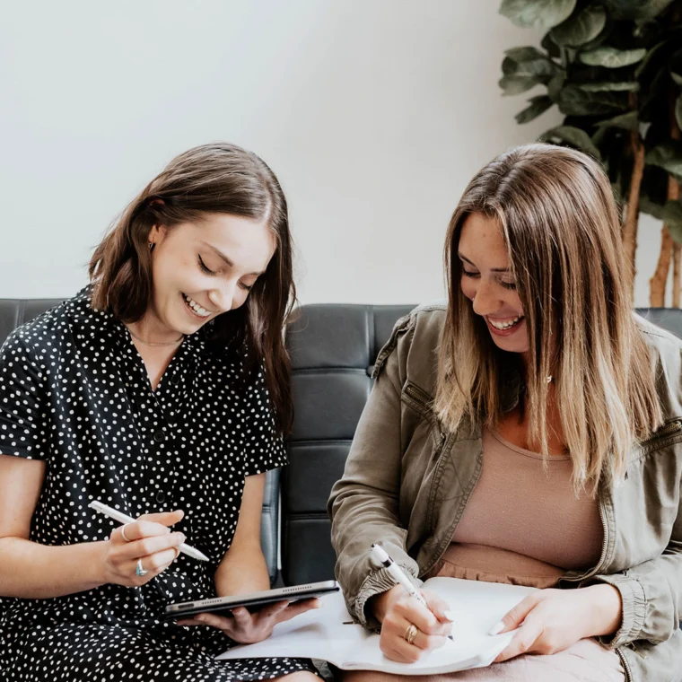 Two women sitting on a couch, smiling and writing notes. One holds a tablet with a stylus, while the other writes on a notepad. There's a plant in the background.