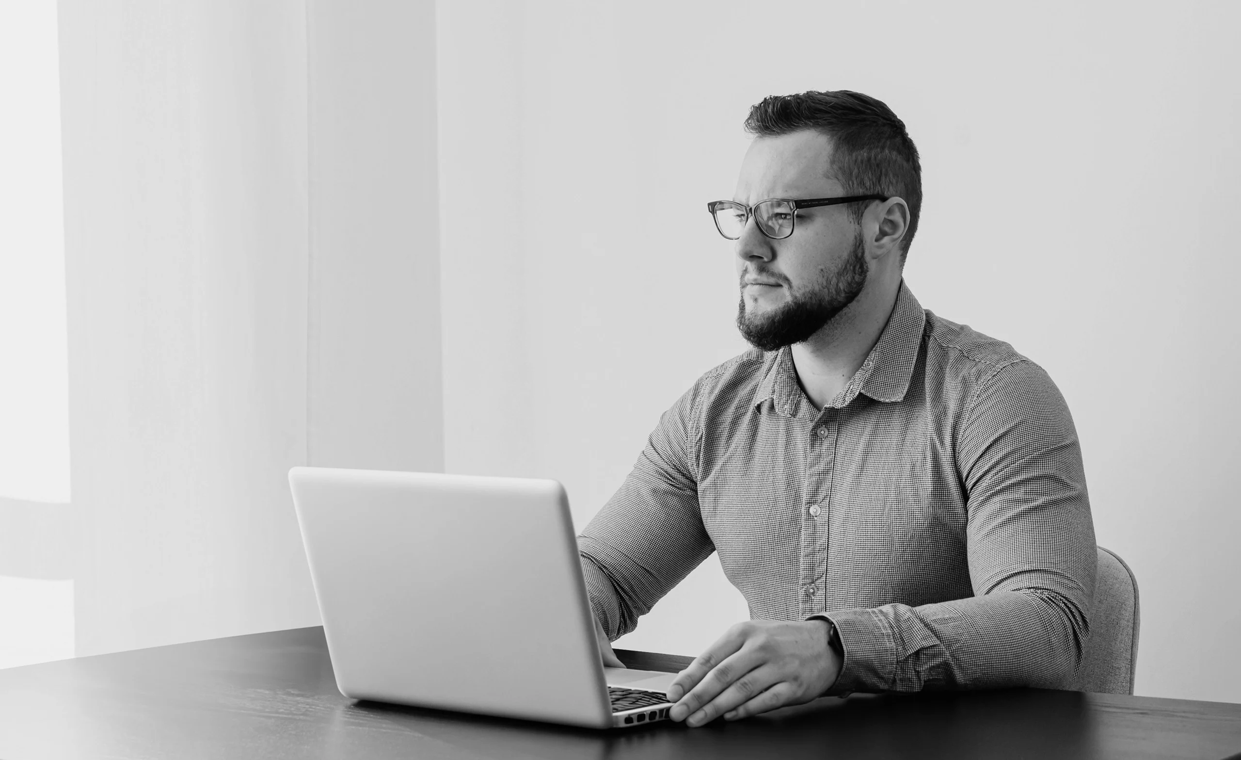 A man with glasses works on a laptop at a table, looking focused. The setting is a bright room with large windows. Black and white image.