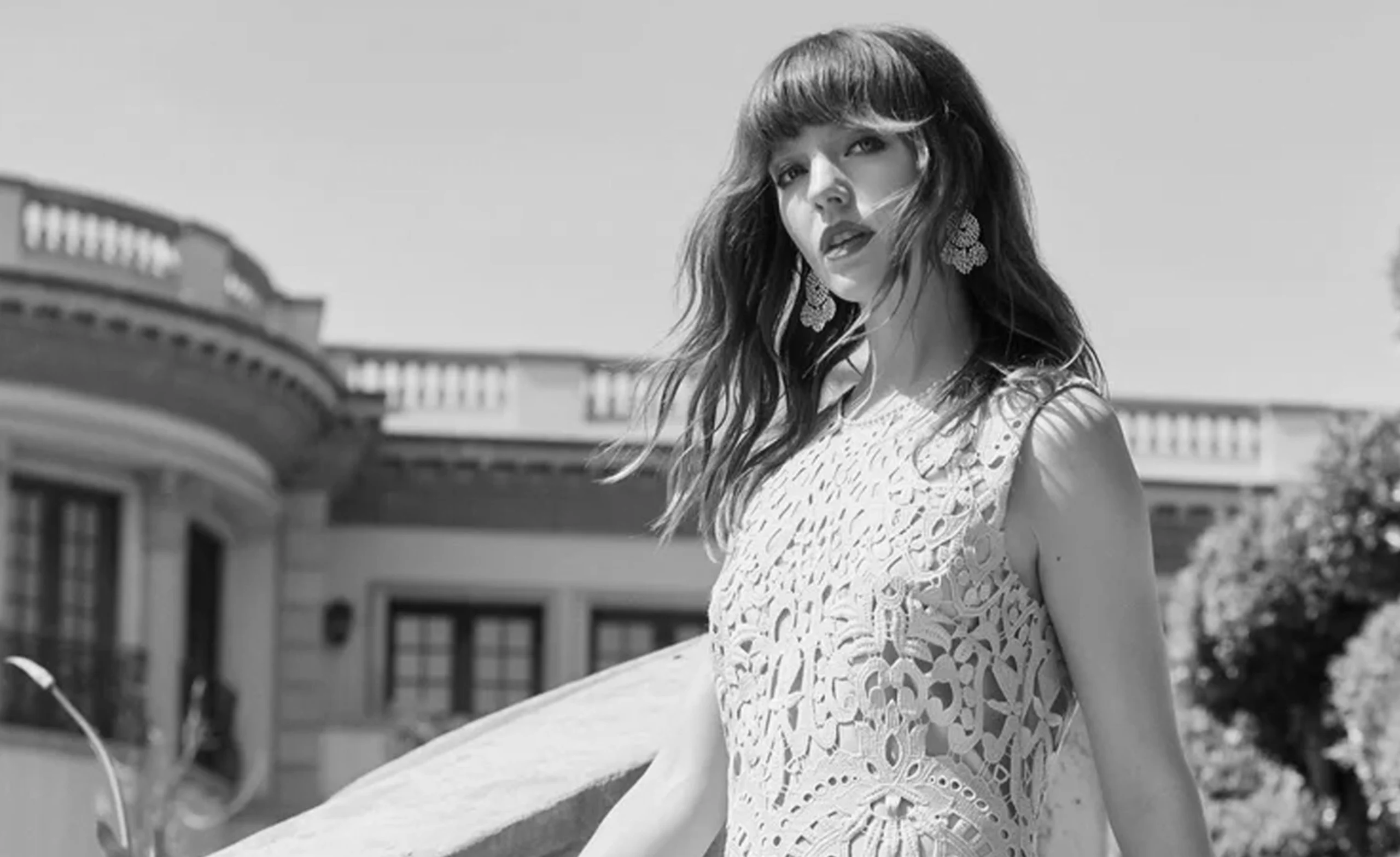 Woman in a lace dress stands outdoors with a historic building in the background, looking at the camera.