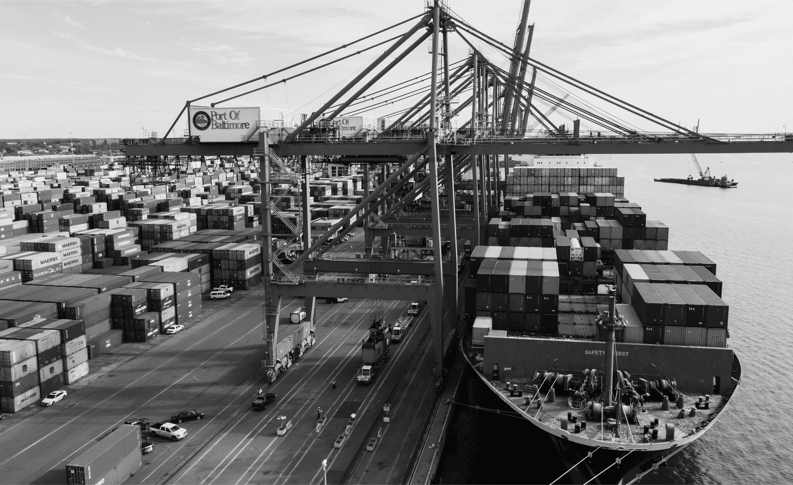 Black and white image of a busy port with cranes loading containers onto a large cargo ship. Multiple stacks of containers and several vehicles are visible on the docks.