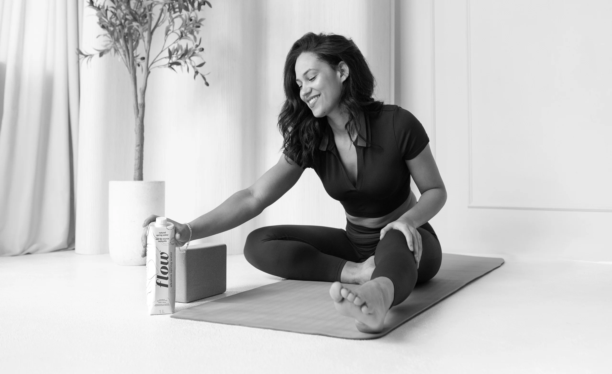 A woman sitting on a yoga mat reaches for a can labeled "Flow" beside a yoga block. A plant is in the background.