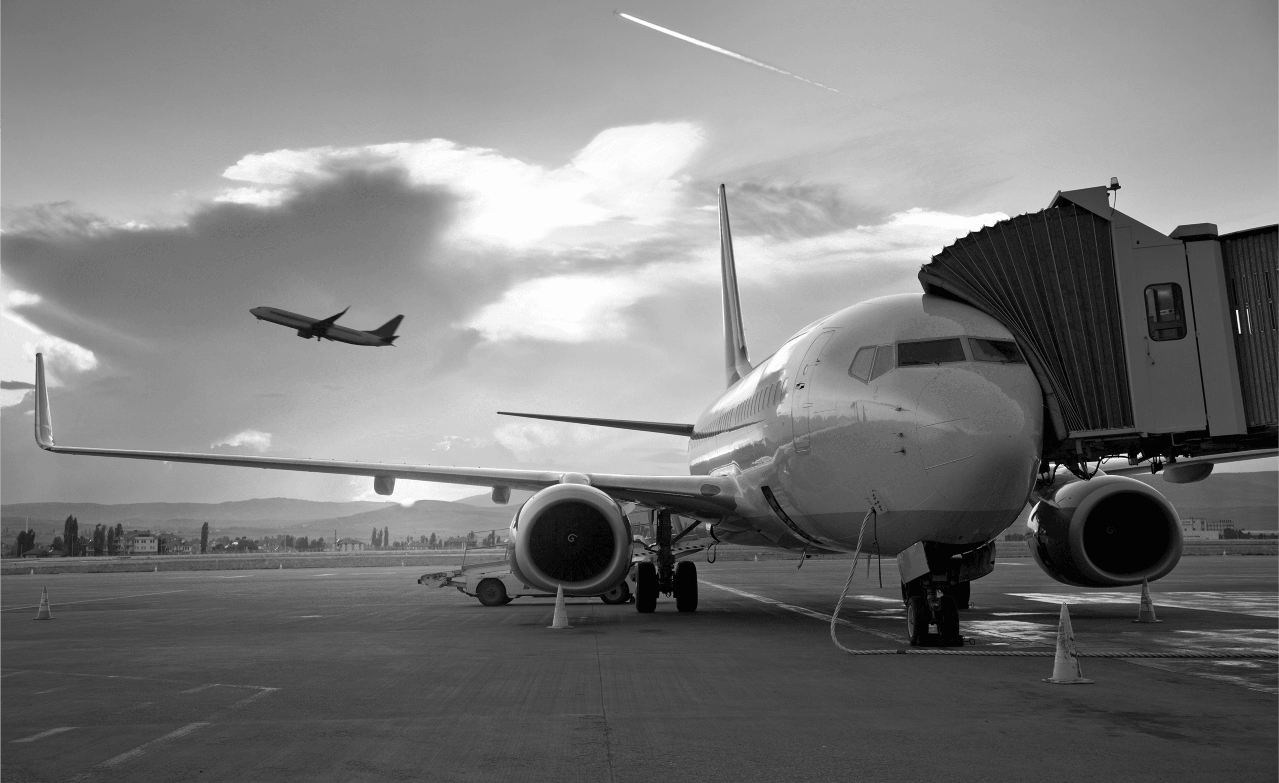 Passenger airplane connected to a jet bridge on the tarmac, with another plane taking off in the background under a partly cloudy sky.