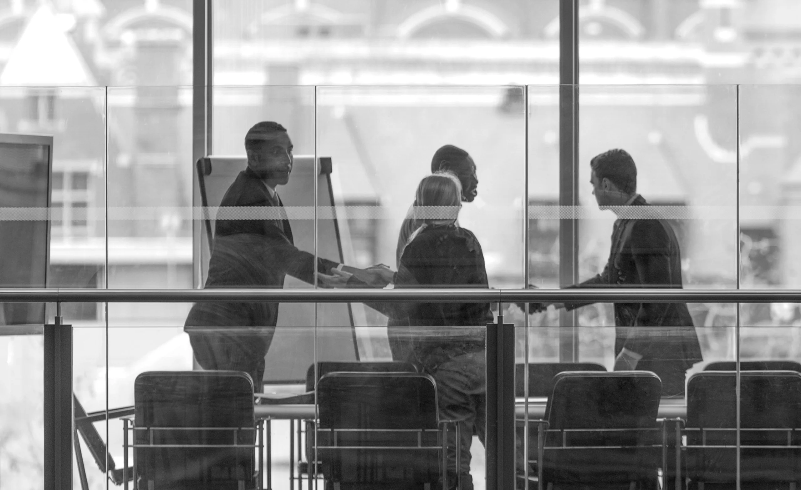 Four people in business attire are engaged in a discussion inside a glass-walled conference room with a view of buildings outside.