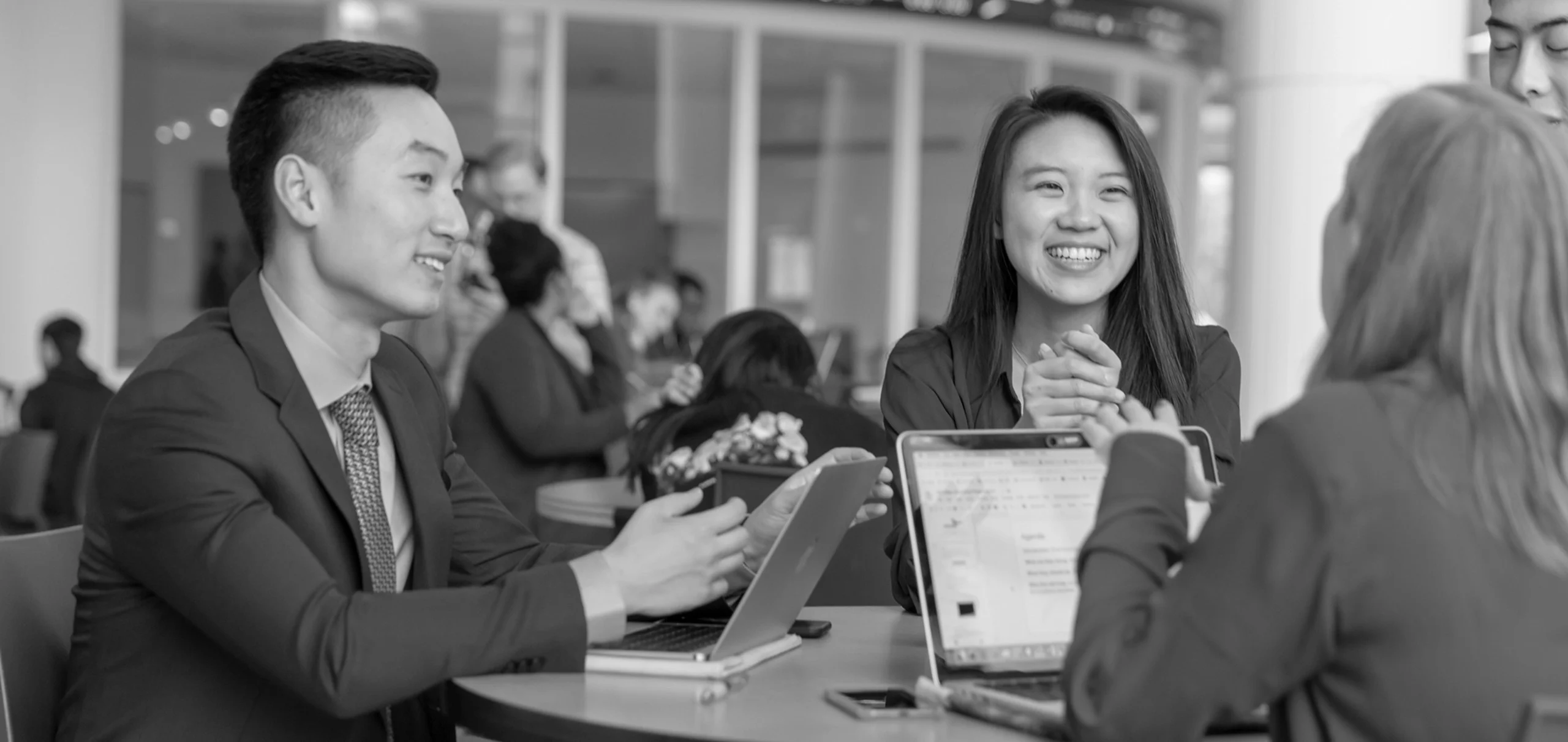 A group of people in business attire sitting at a table with laptops, engaged in conversation and smiling.