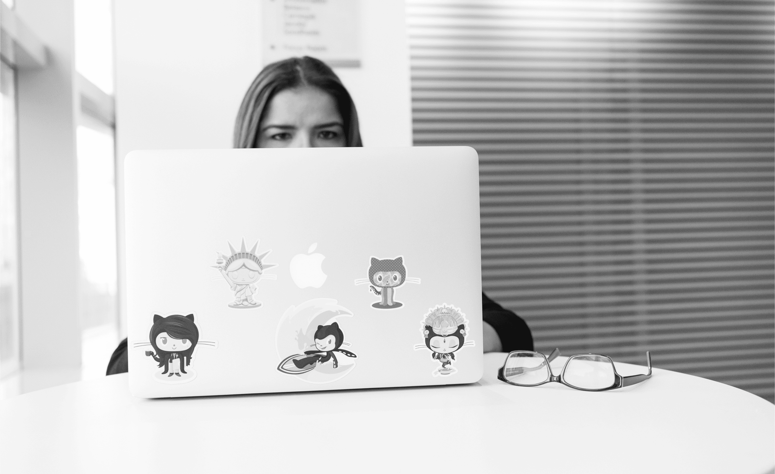 A person sits behind a sticker-covered laptop at a white table. Glasses are placed on the table to the right of the laptop.