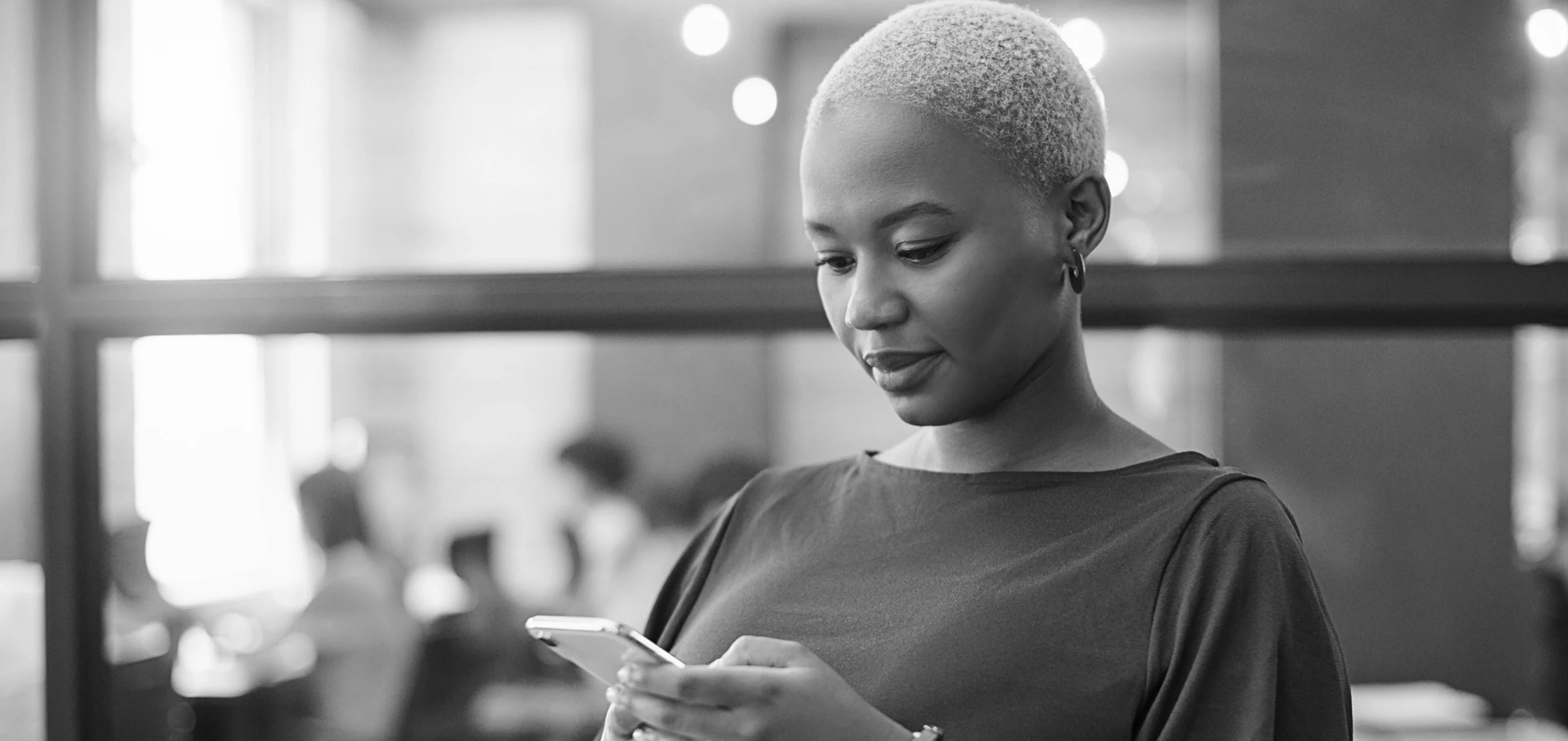 Person with short hair, wearing a dark top, looks at their smartphone. The background shows an office environment with blurred figures.
