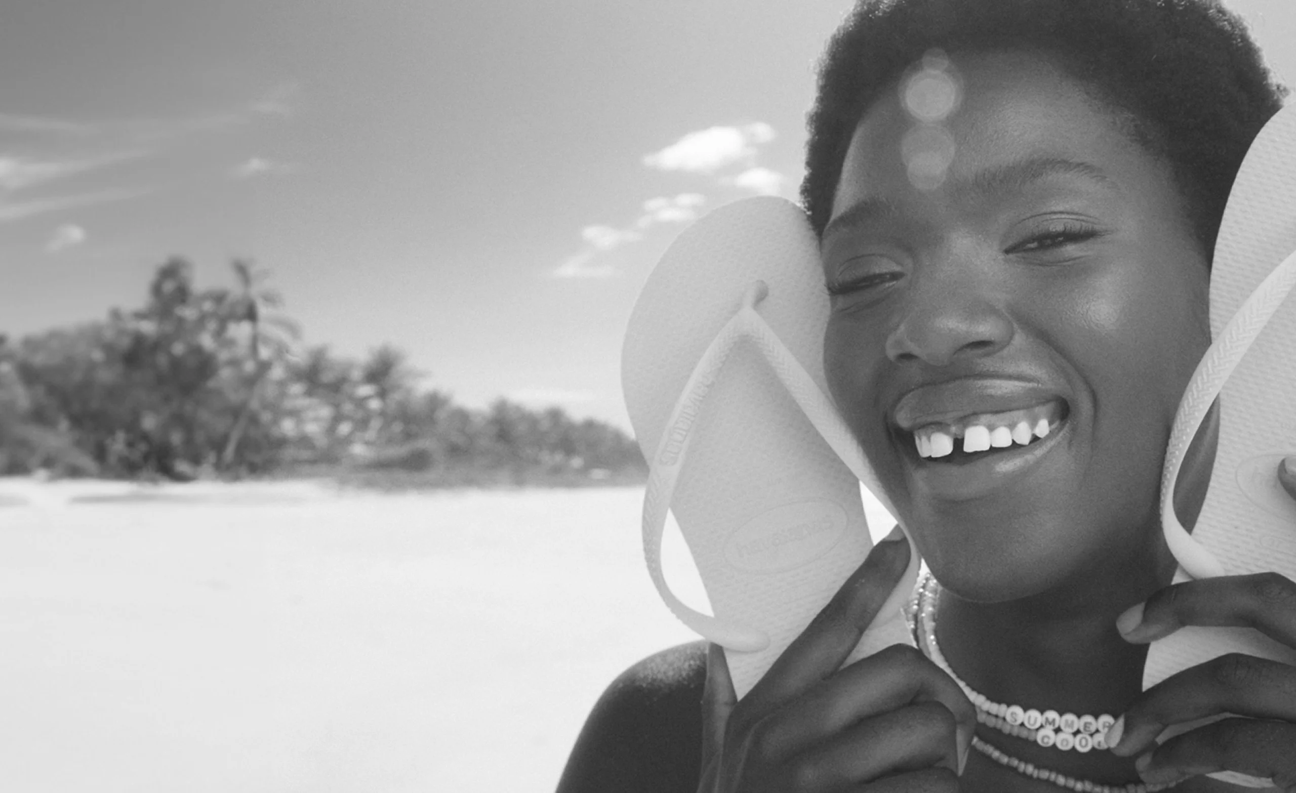 Person smiling on the beach, holding flip-flops by their face, wearing a necklace.