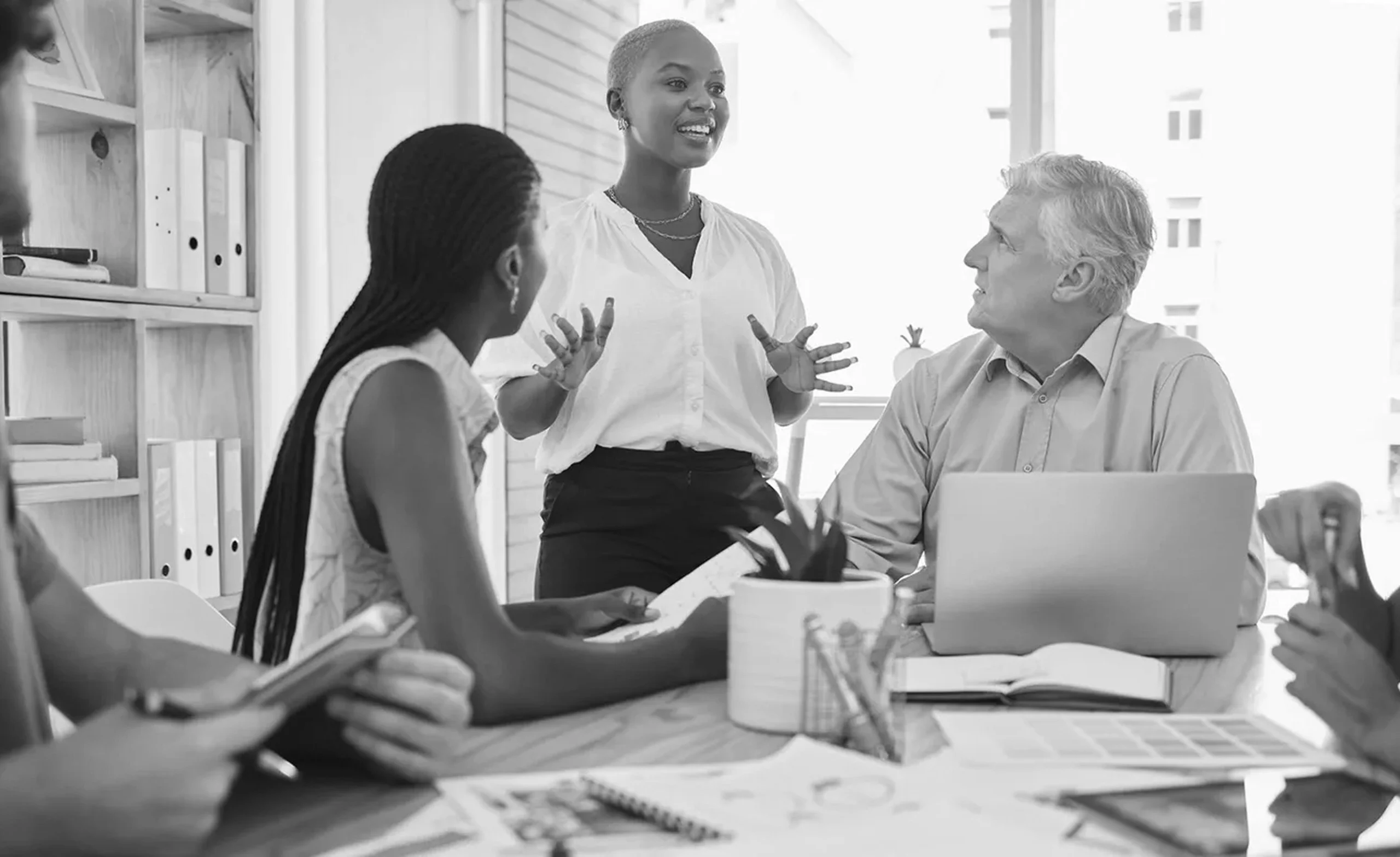 Three people in a business meeting. A woman is standing and speaking, while a man and another woman are seated and listening. Papers and a laptop are on the table. Black and white image.