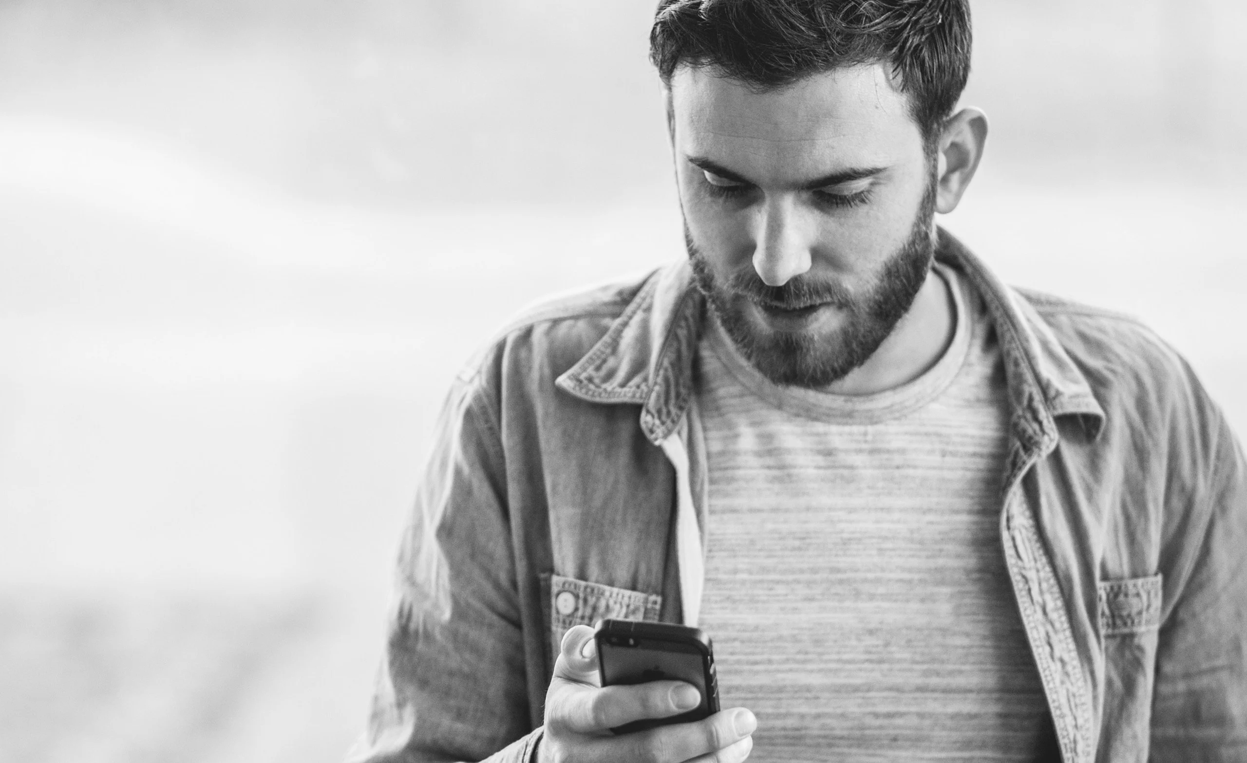 A man with a beard and wearing a jacket looks at his phone screen in a grayscale image.