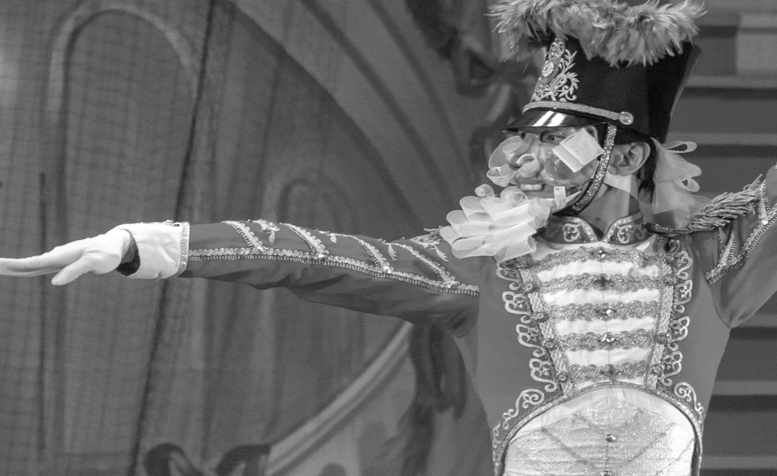 Performer in an ornate, military-style costume with a feathered hat and mask, extending one arm outward against a stage backdrop.