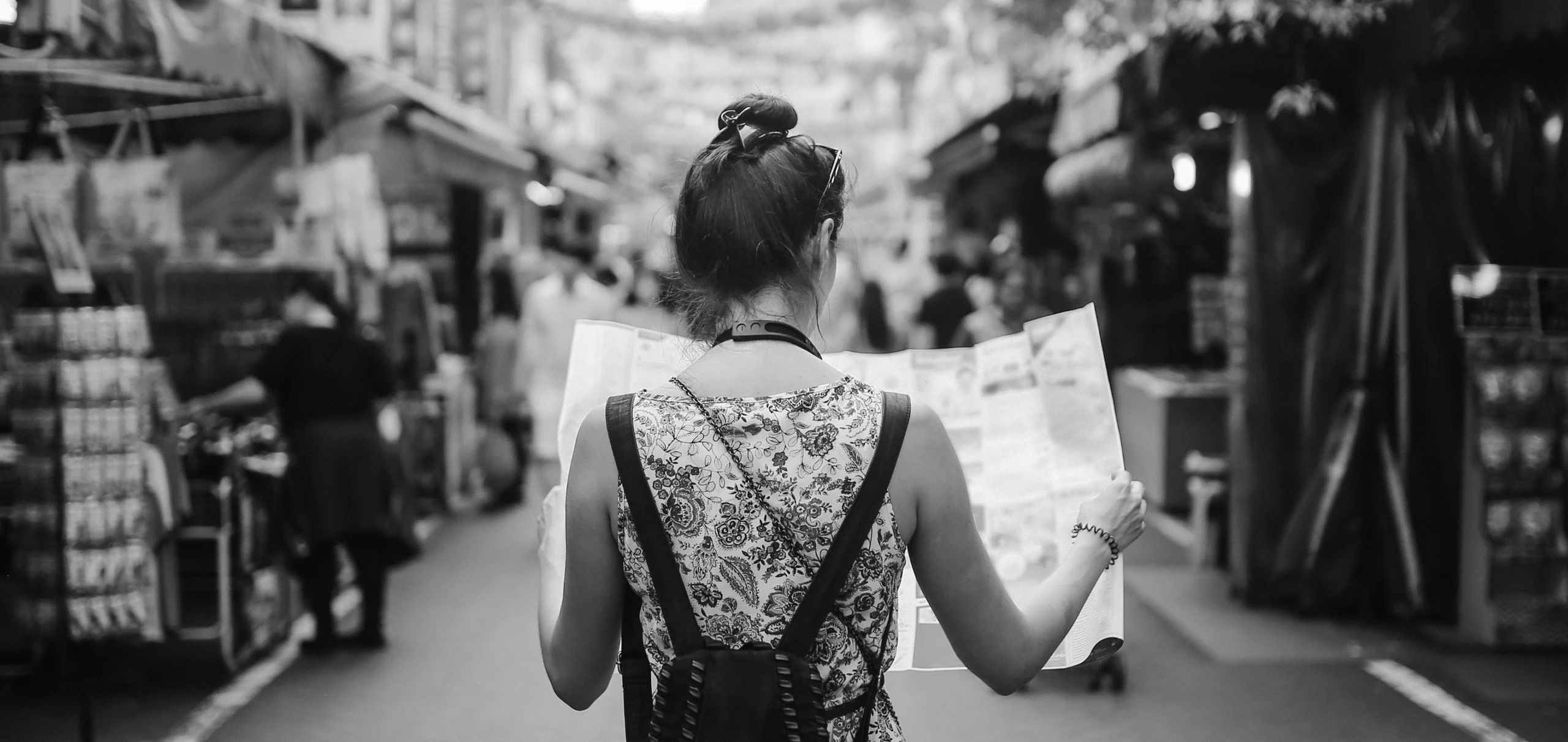 A woman stands in a busy outdoor market, back to the camera, holding a map. She wears a floral dress and has a backpack. Black and white photo.