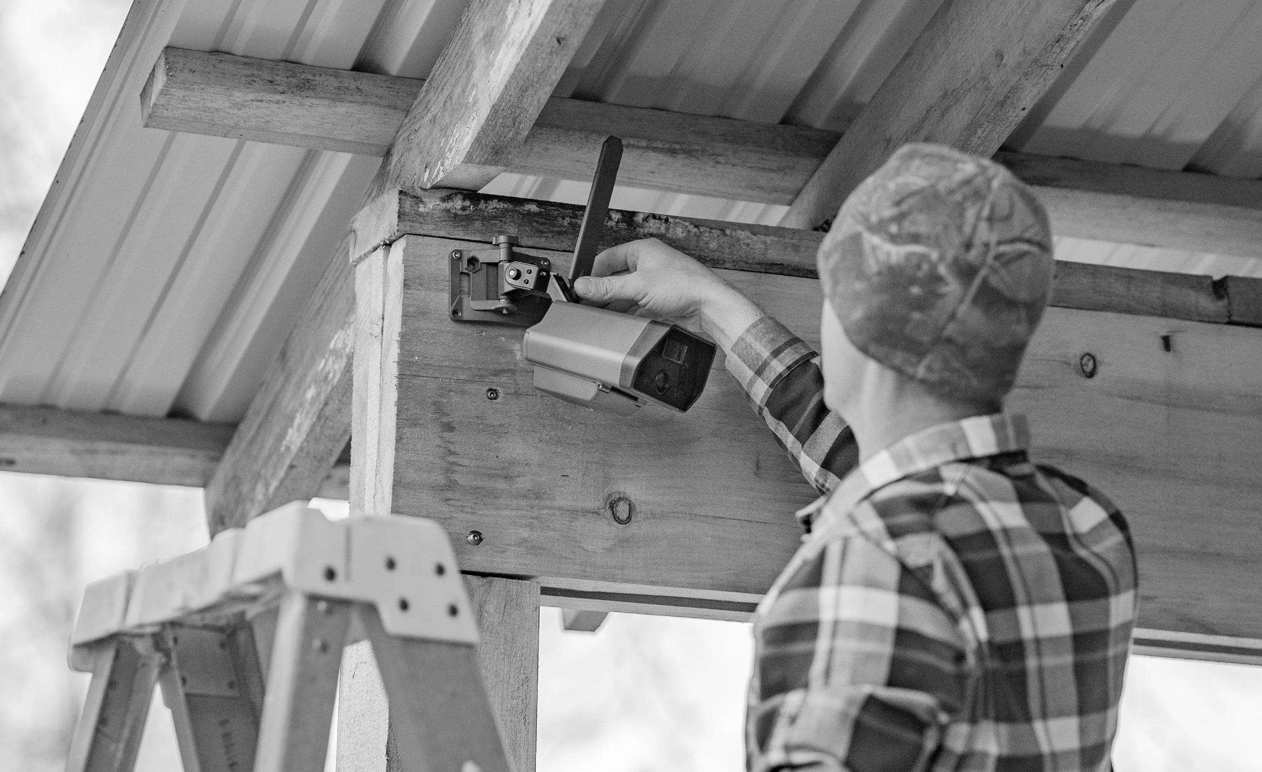 A person in a plaid shirt and cap installs a security camera on a wooden beam, standing on a ladder beneath a corrugated metal roof.