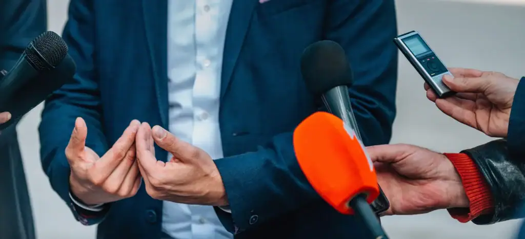 A person speaking to people with multiple microphones in front.