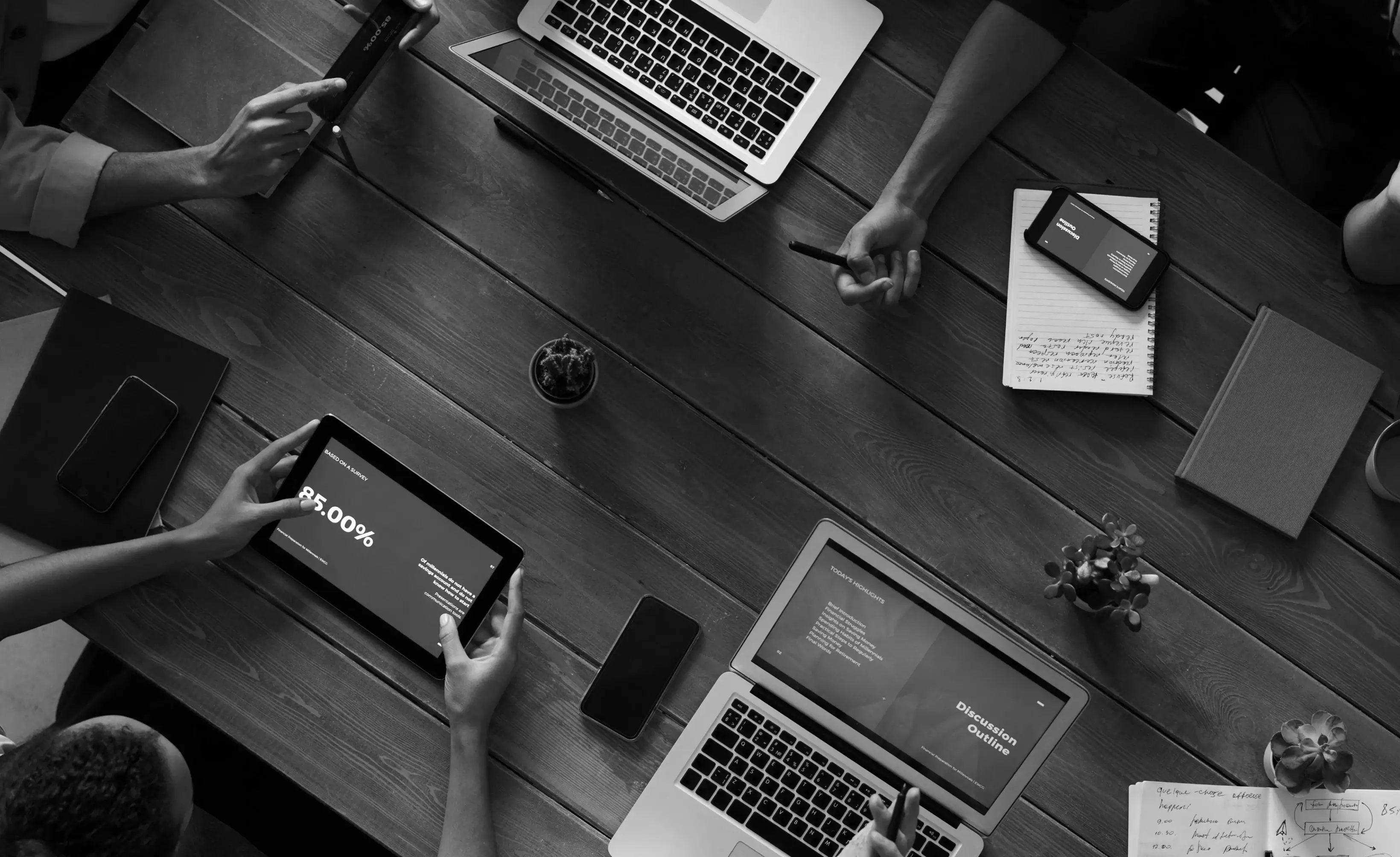 Four people work at a wooden table with laptops, a tablet showing data, a phone, notebooks, pens, and small plants visible, viewed from above.