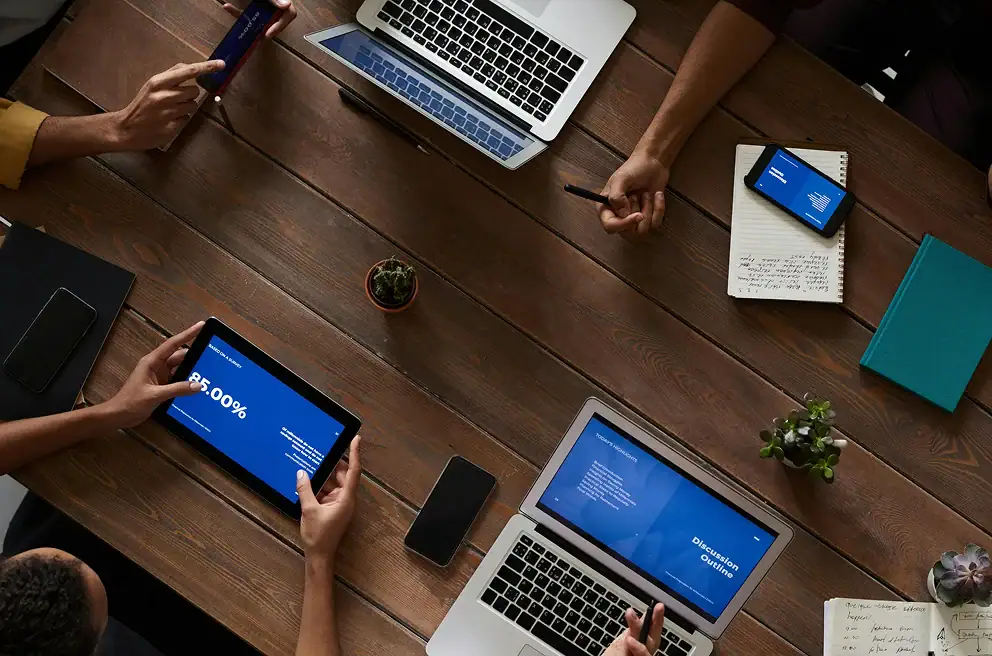 Four people sit at a wooden table with laptops, tablets, a notebook, and a smartphone, appearing to collaborate on a project or presentation.