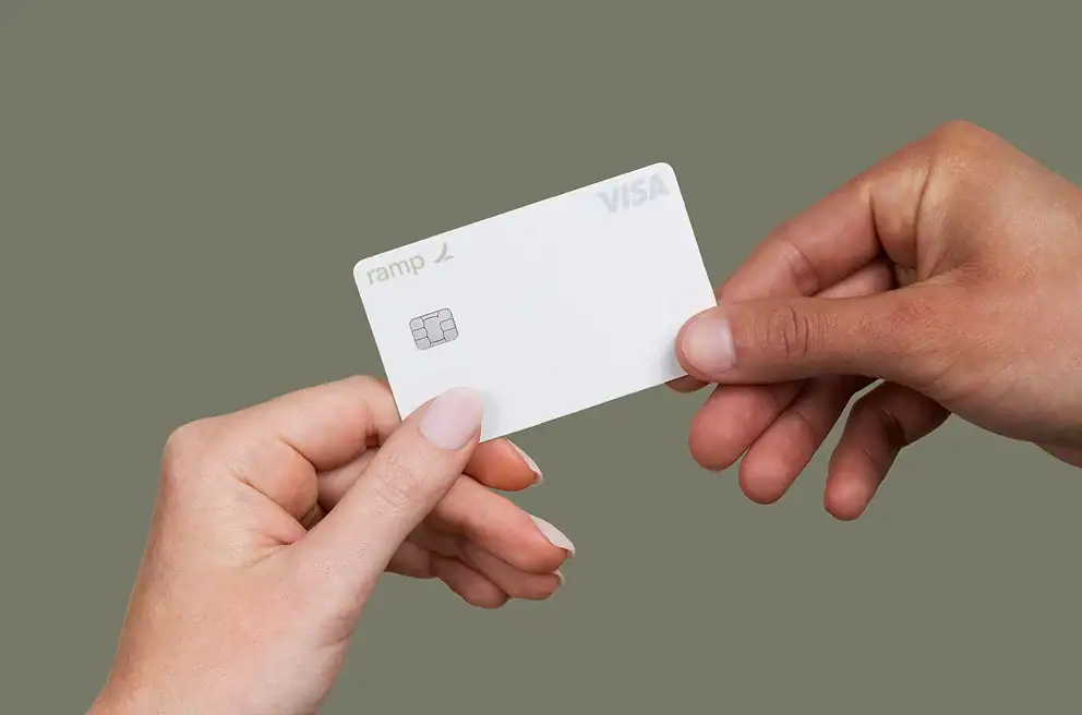 Two hands exchanging a white Ramp VISA credit card against a plain, muted background.