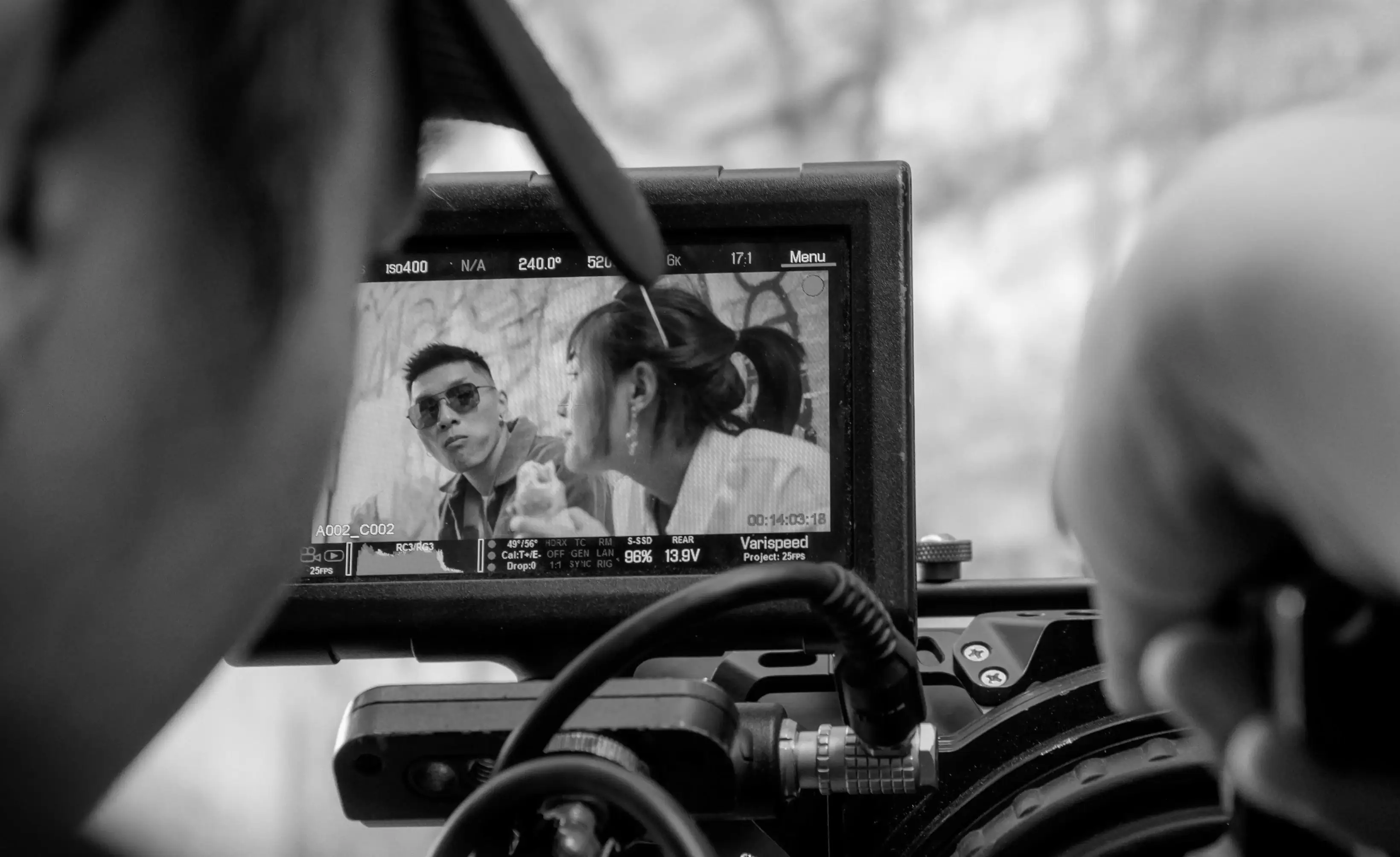 A camera monitor displays a black and white scene of two people talking, as seen from behind the camera on a film set.