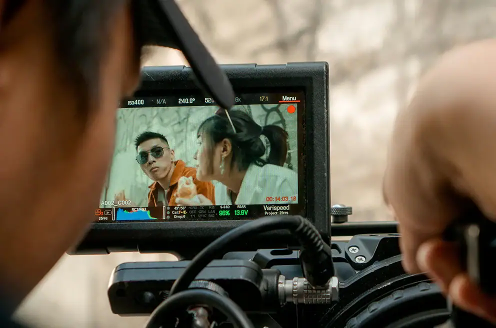 A camera monitor displays two people in conversation during a film shoot, with a crew member adjusting the camera in the foreground.