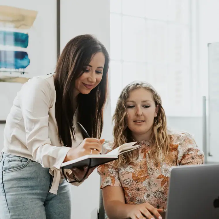 Two women work together at a desk; one writes in a notebook while the other looks at a laptop screen.