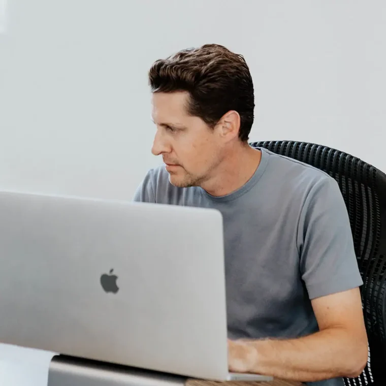 A man in a gray t-shirt sits at a desk, working on a laptop with an Apple logo. He faces left, focused on the screen, in a minimalist office setting.